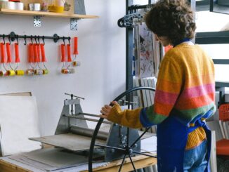 Side view of anonymous female master working at workbench with printing press with wheel in modern art studio with rollers